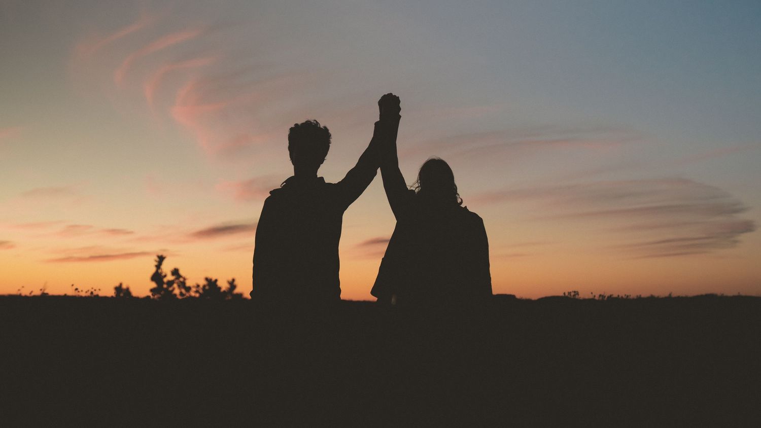 Silhouetted figures stand together against a vibrant orange and purple sunset sky on the horizon.