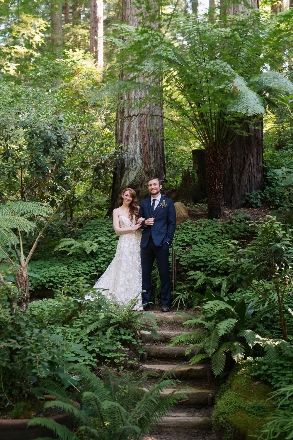 A romantic wedding couple poses along mossy stone steps in a lush forest setting among redwood trees and ferns.