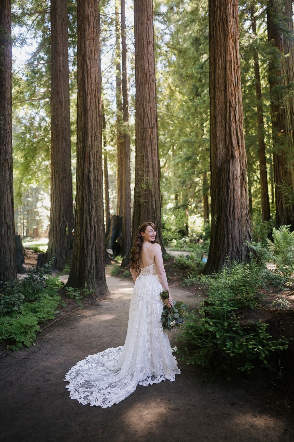 A bride in a white lace wedding dress with a long train stands on a forest path among towering redwood trees.