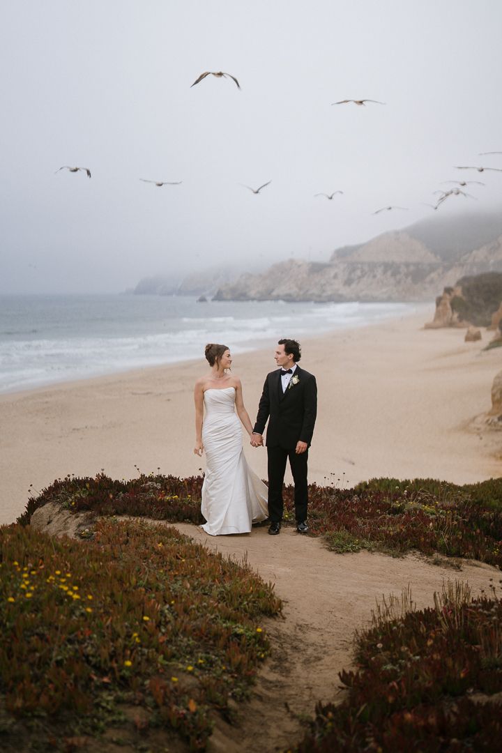 A couple in wedding attire stands on a coastal path overlooking a misty beach with seagulls flying overhead.