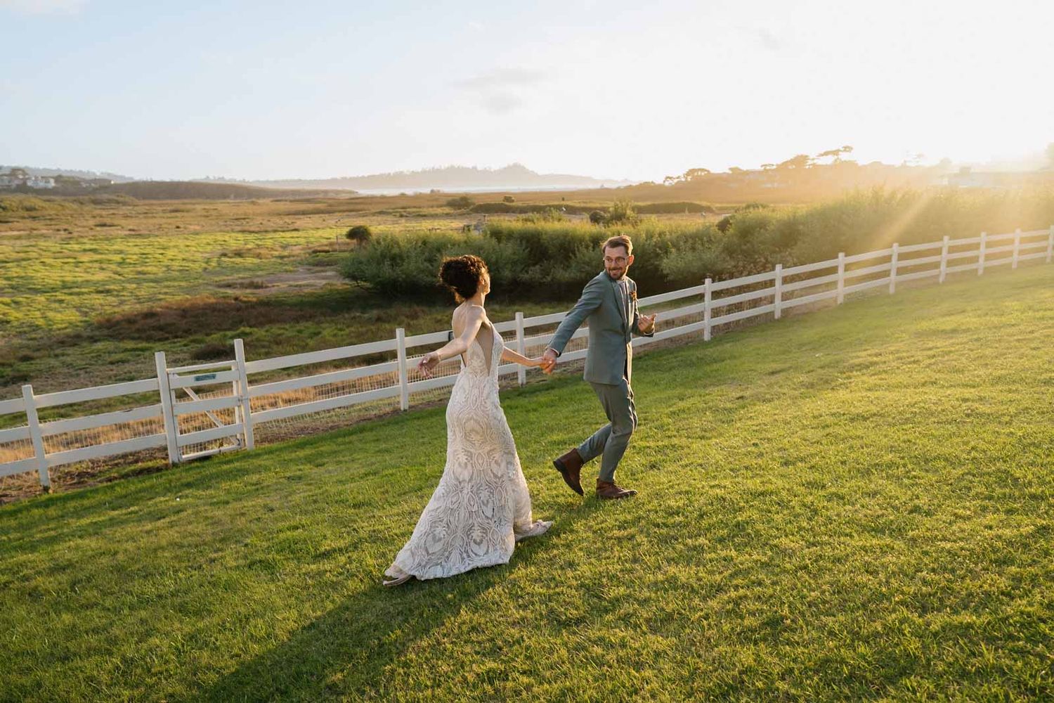 Two people run hand in hand across a sunlit meadow with mountains in the background at sunset at mission ranch wedding venue in Carmel.