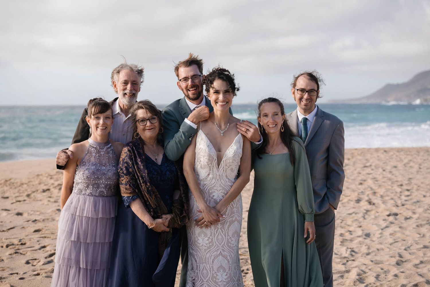A group of people in formal attire gather on a sandy beach with ocean waves and mountains in the background.