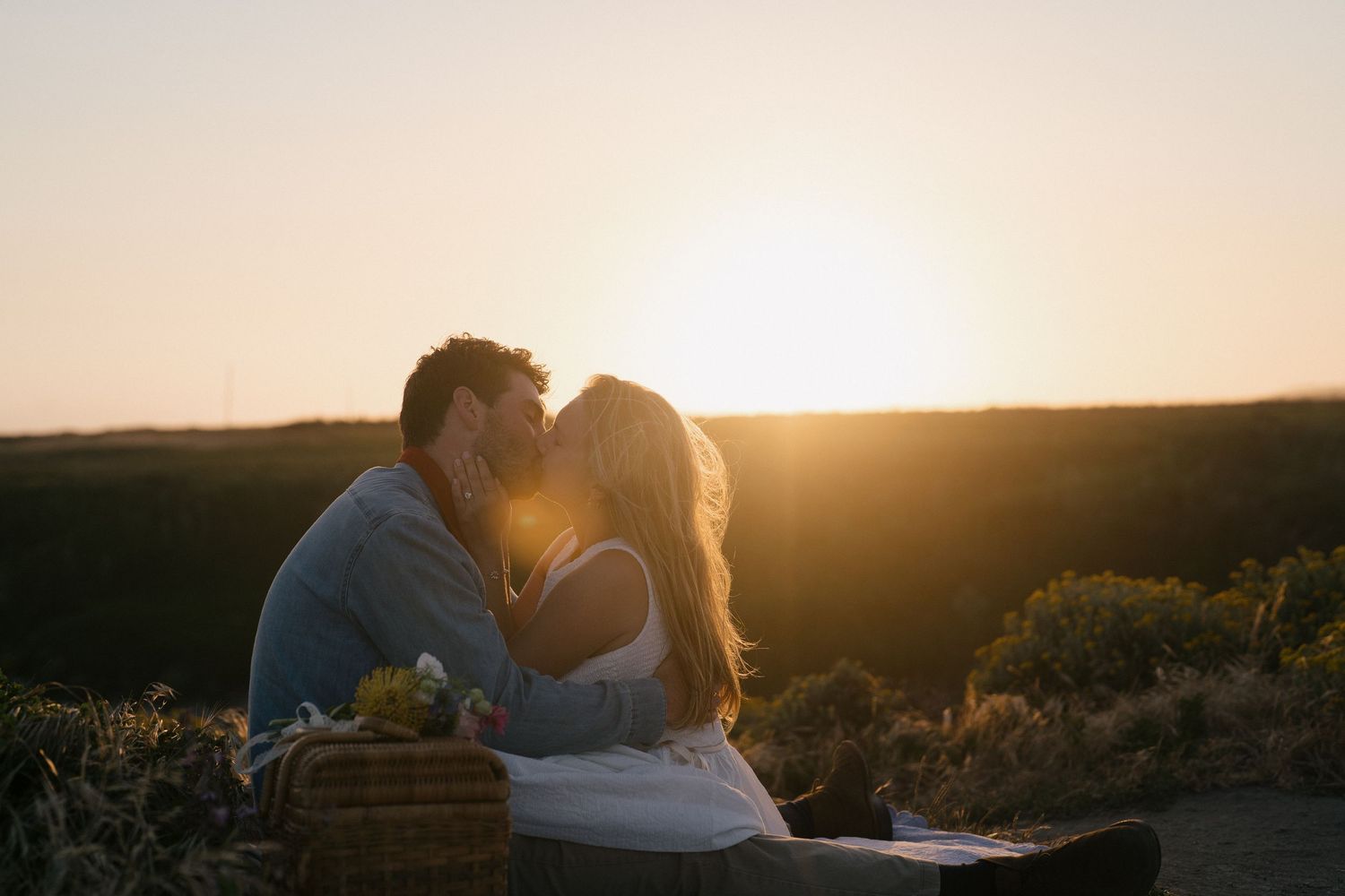 A romantic couple sits on a picnic blanket sharing an intimate moment as the sun sets behind them in a golden field.