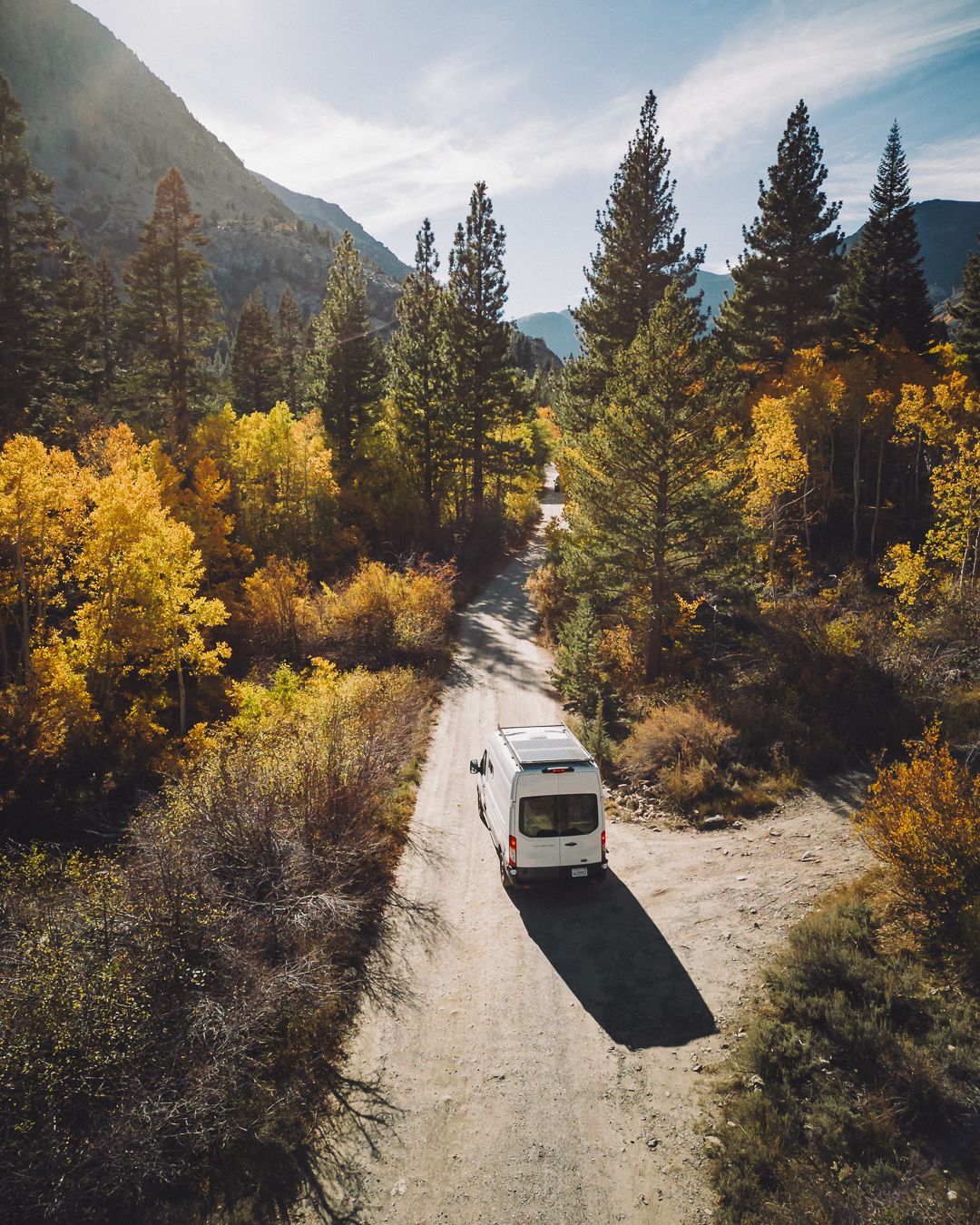 An RV drives along a winding dirt mountain road surrounded by vibrant autumn trees and rugged peaks.