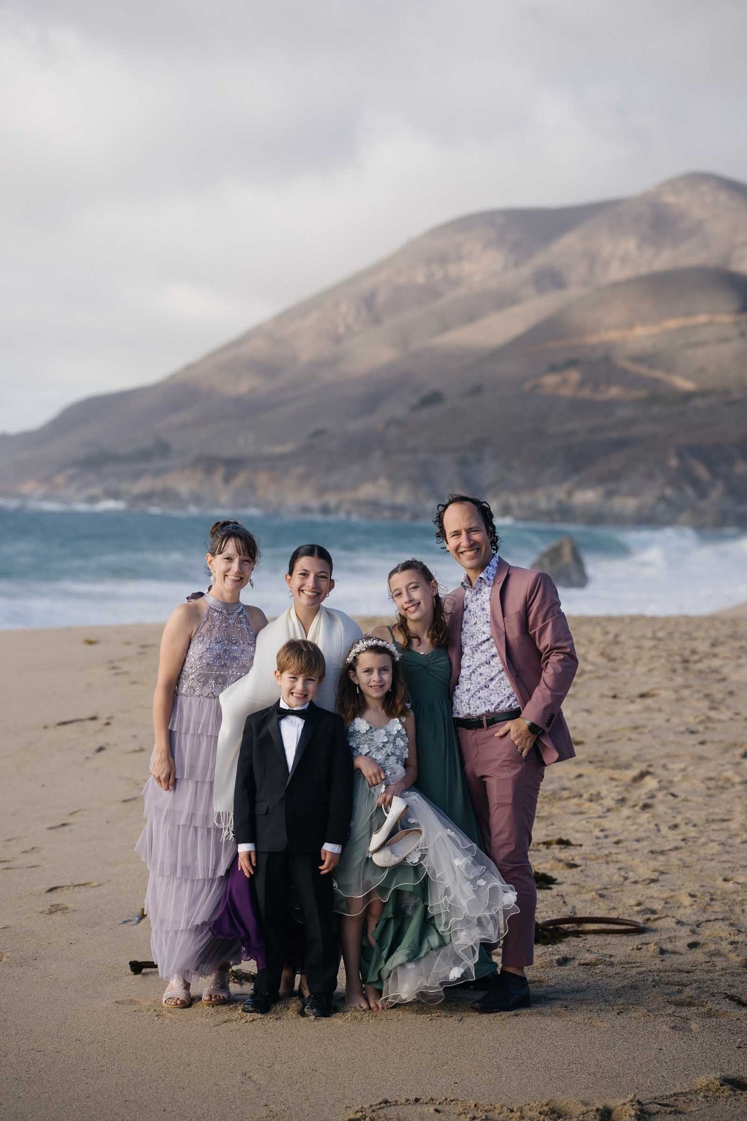A group poses together on a scenic beach with mountains in the background wearing formal and semi-formal attire.