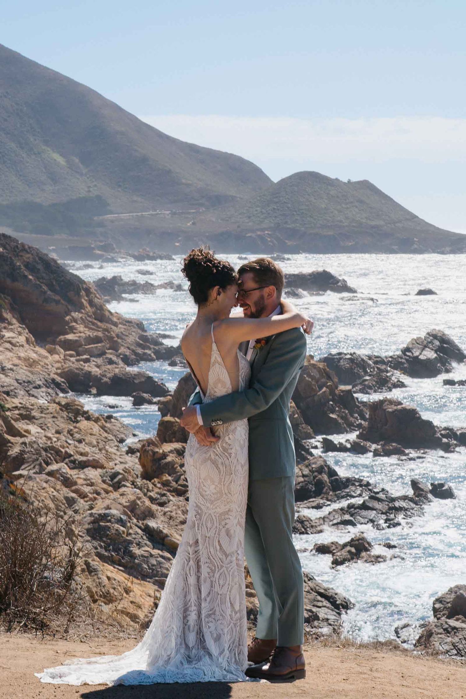 A couple embraces on a cliff overlooking a dramatic rocky coastline with crashing waves and mountains in the background.