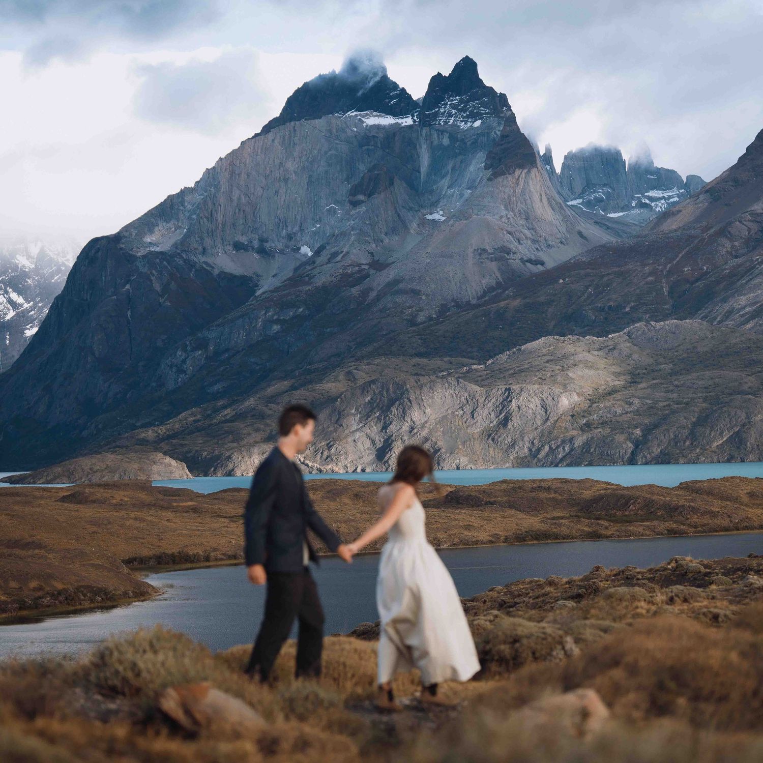 A couple dances on rugged terrain with dramatic mountain peaks and a serene lake in Torres del Paine National Park, Chile.