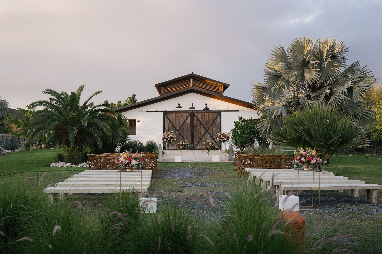 A panoramic view of a ranch property with horses grazing near white fences and palm trees at sunset.