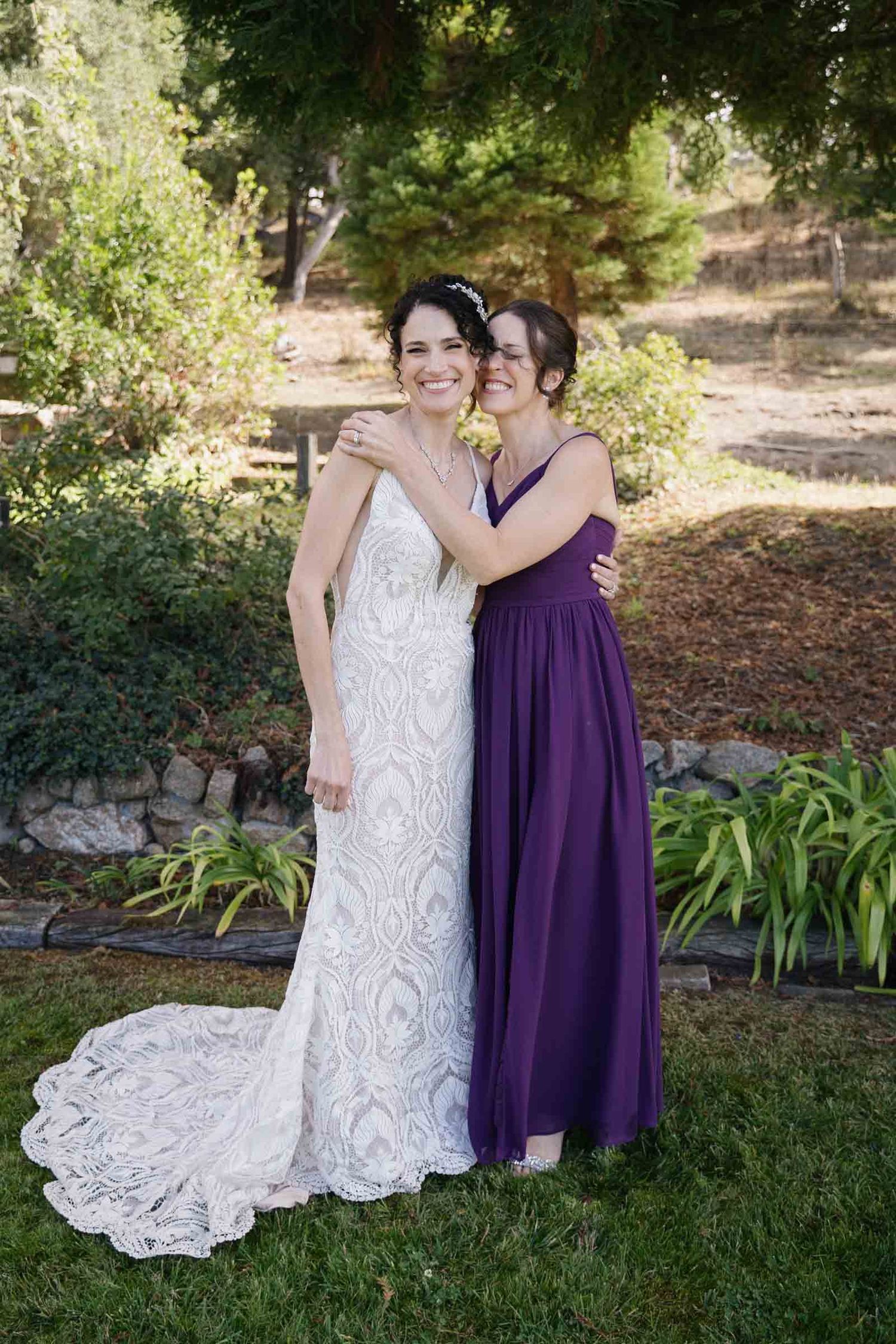 Two people pose together outdoors, one wearing a white lace dress and the other in a deep purple dress.