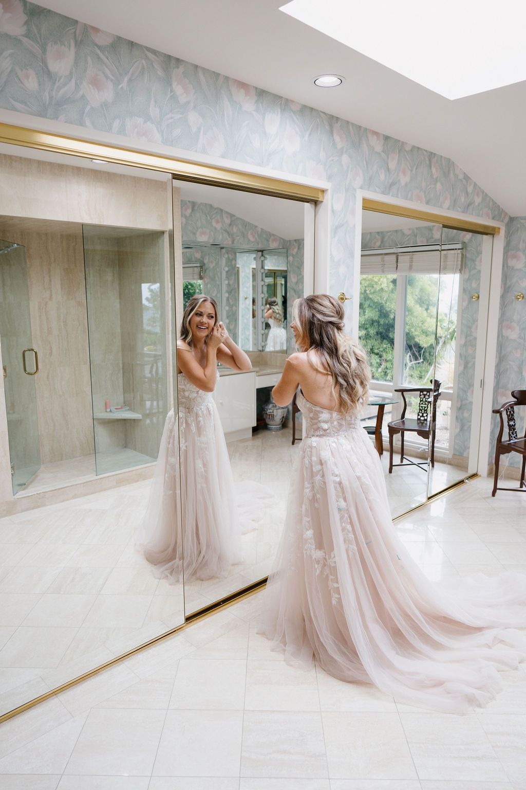 A beautiful wedding dress with delicate lace details and flowing tulle skirts reflects elegantly in a gold-framed mirror.