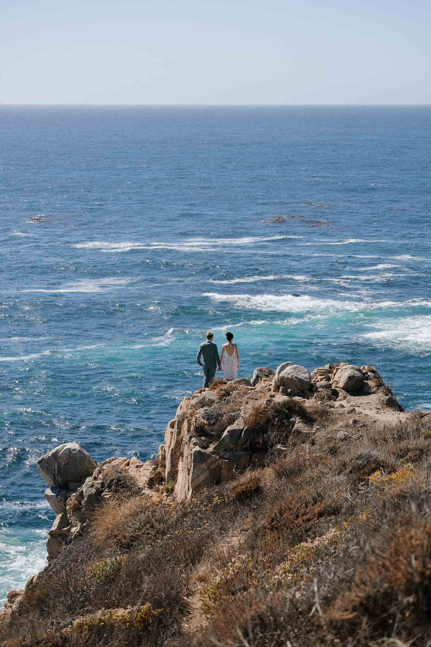 People stand on a rocky coastal cliff overlooking the blue Pacific Ocean waves crashing against the shore.
