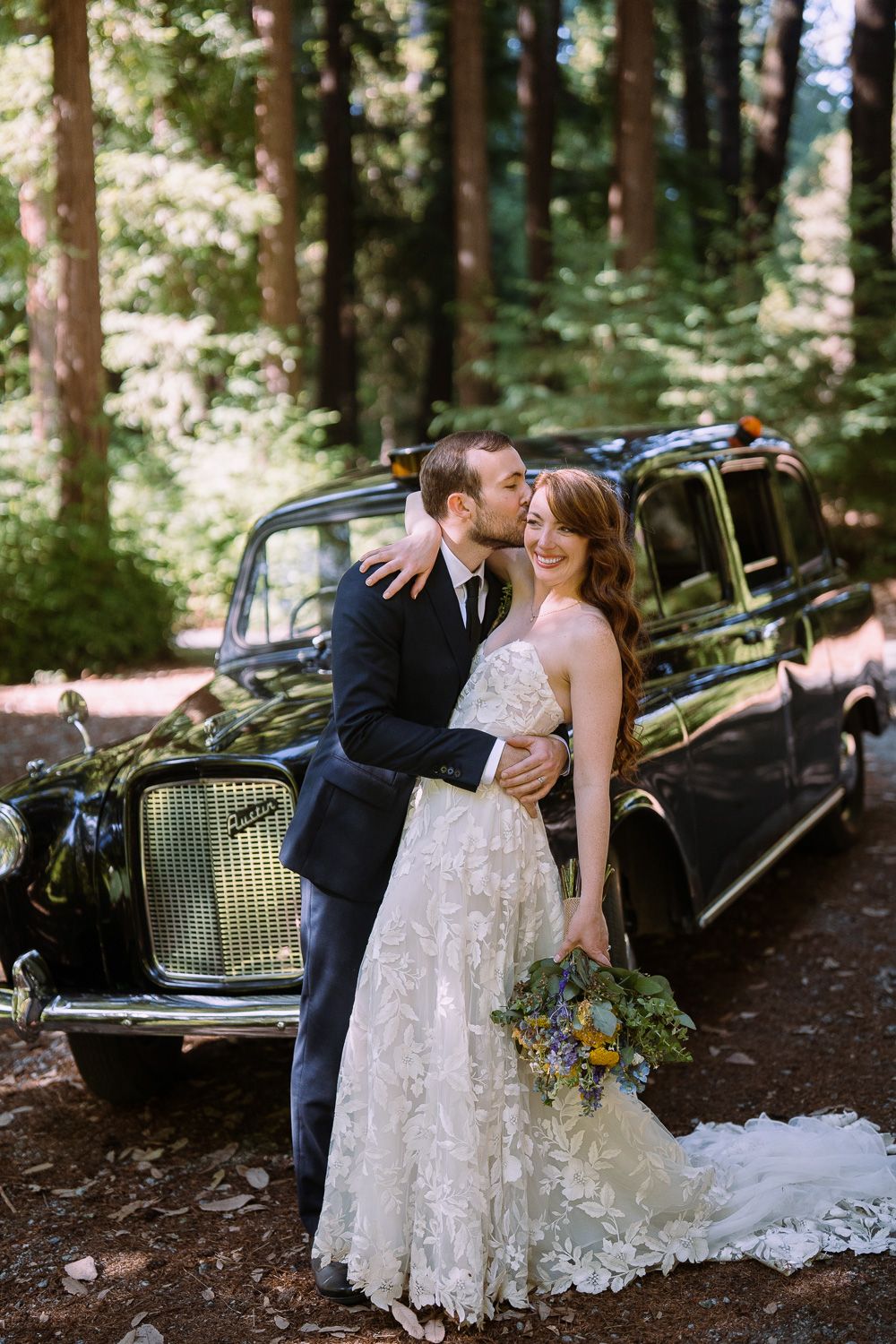 A romantic wedding couple embraces next to a vintage black car in a serene forest setting with dappled sunlight.