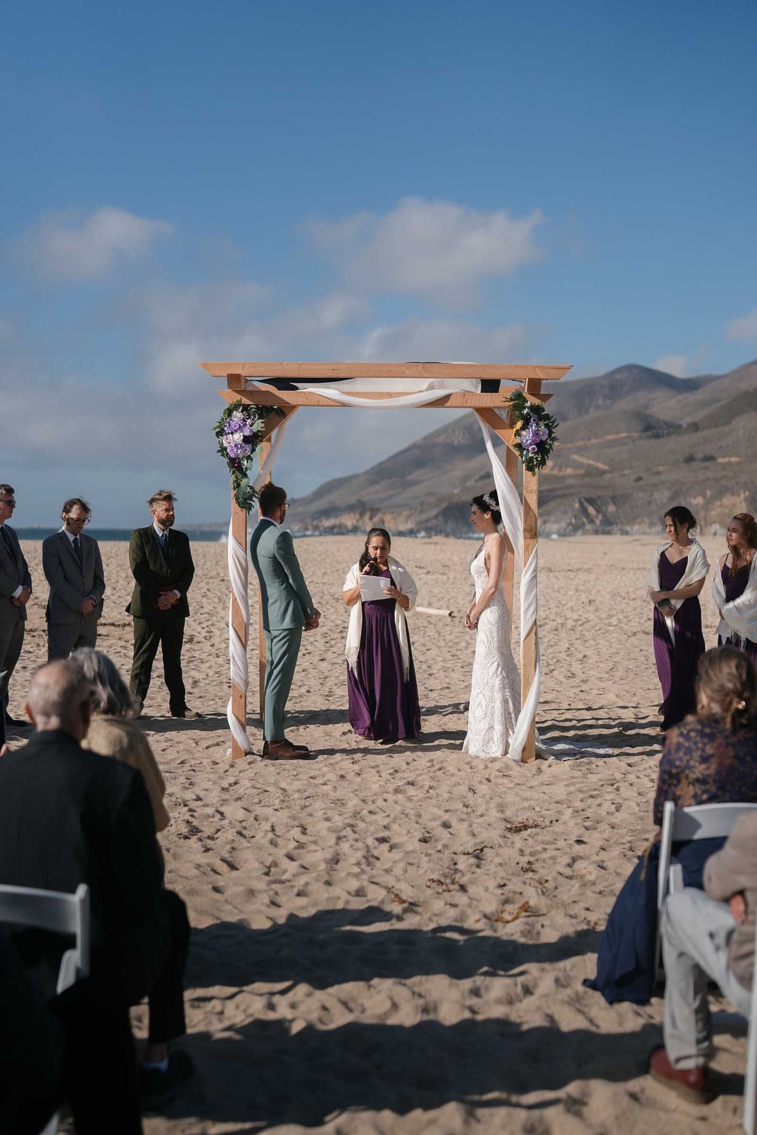 A crowd of people holding purple parasols gather outdoors for a wedding ceremony at garrapata beach in big sur on a sunny day.