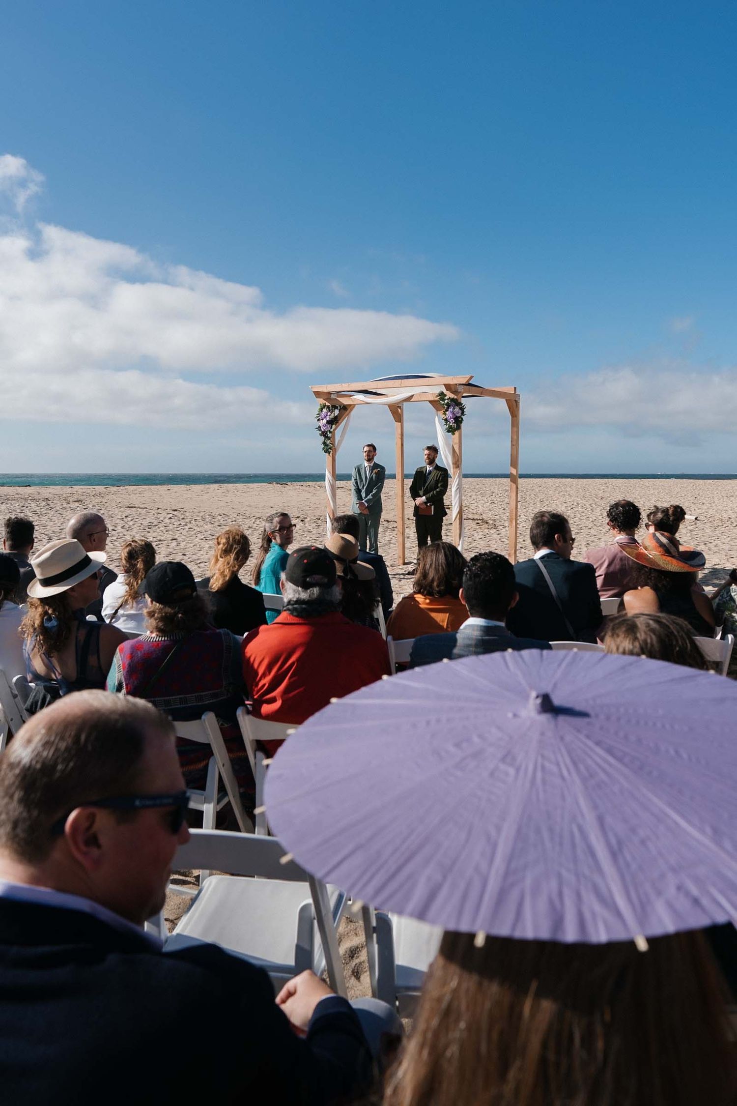 A crowd of people holding purple parasols gather outdoors for a wedding ceremony at garrapata beach in big sur on a sunny day.