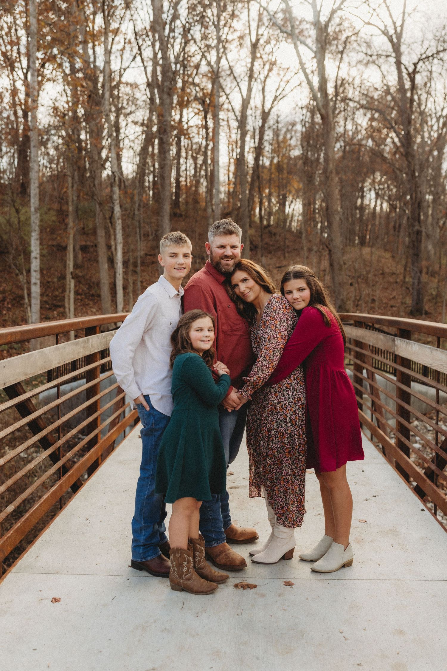 Family of five posing together on a wooden bridge surrounded by autumn trees, wearing coordinated red and green outfits.
