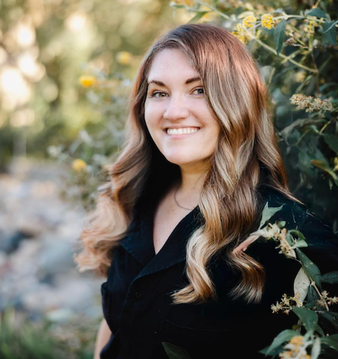 Professional outdoor portrait with long wavy hair, wearing black against blurred yellow flowers and natural foliage.