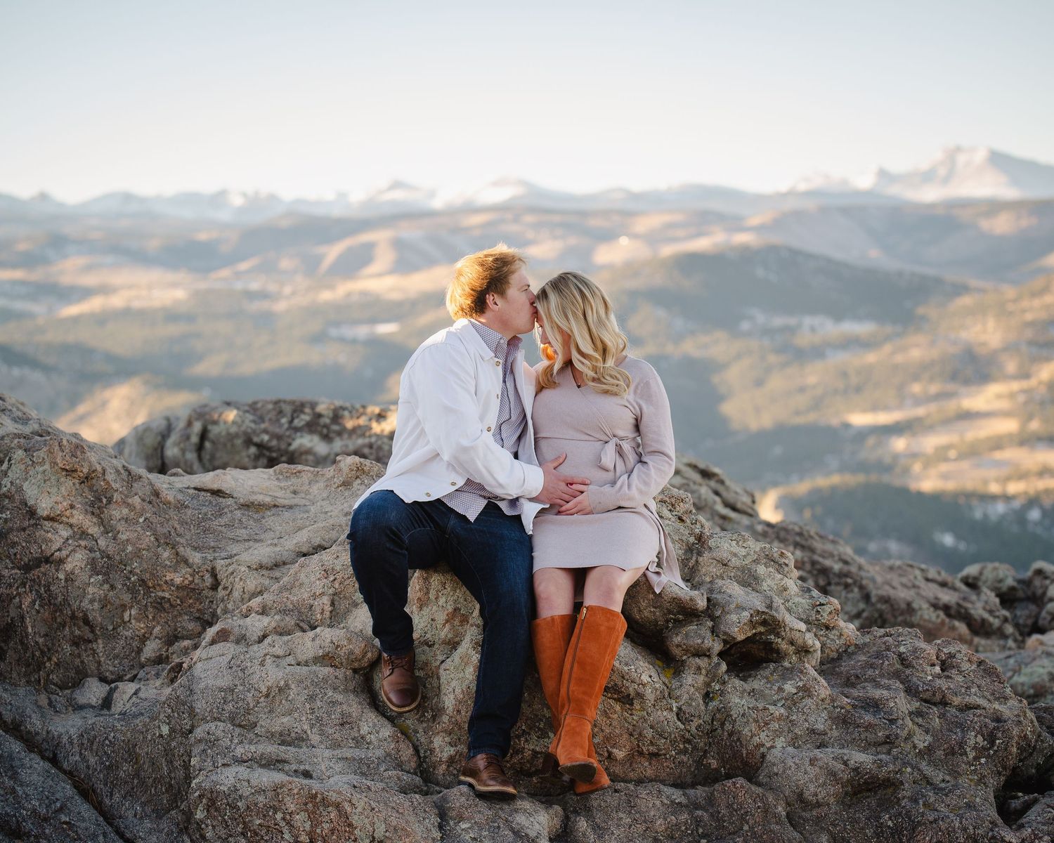 A couple sits together on a rocky mountain overlook during sunset with scenic mountain ranges in the background.