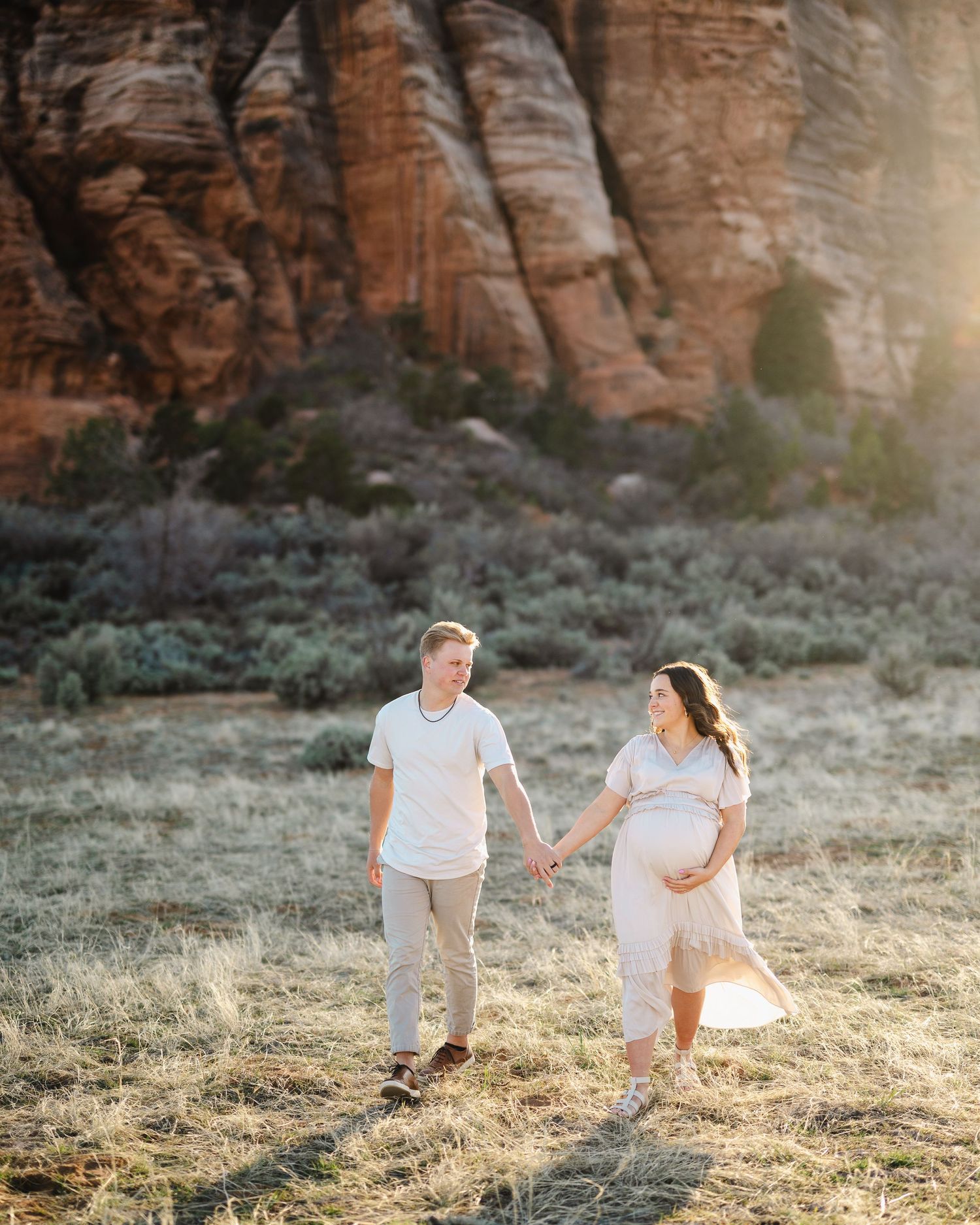 A couple walks hand in hand through a desert landscape with dramatic red rock formations in the background.