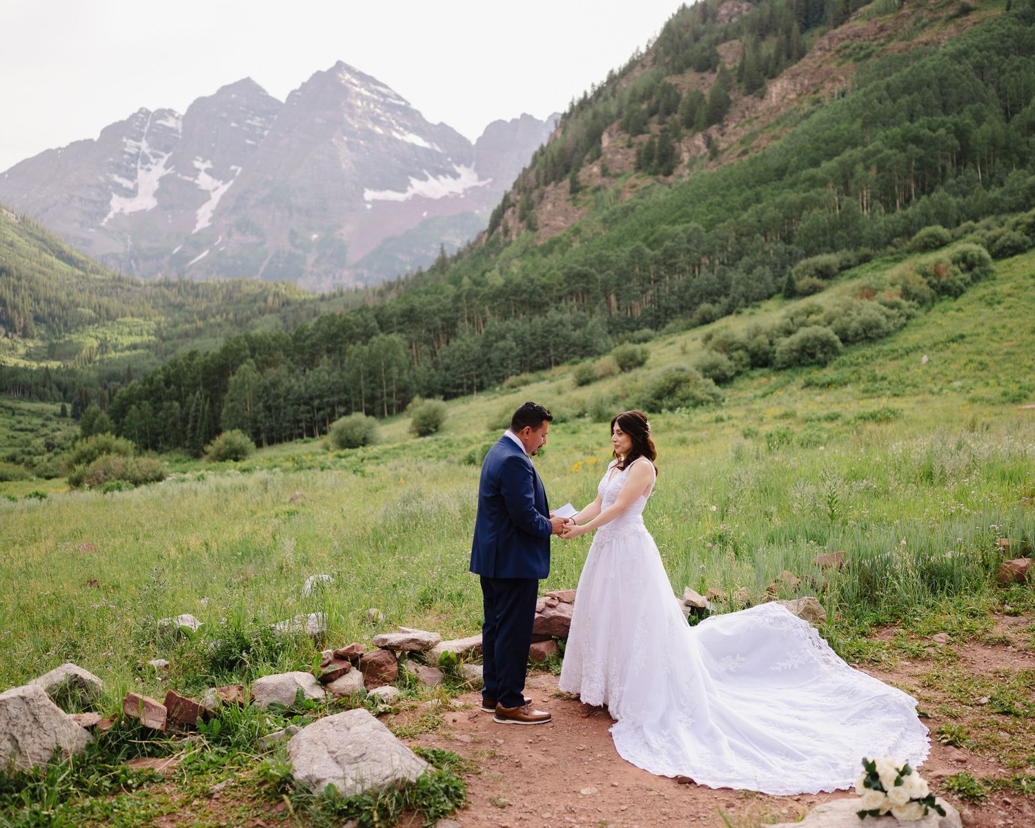 A couple in wedding attire stands near majestic snow-capped mountains with lush green meadows in the background.