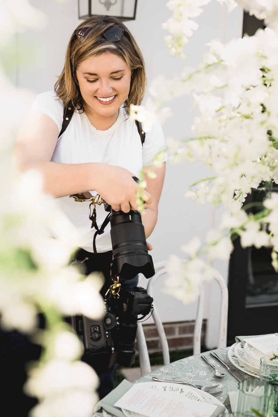 Smiling female photographer holding a DSLR camera surrounded by white floral arrangements at an outdoor event.