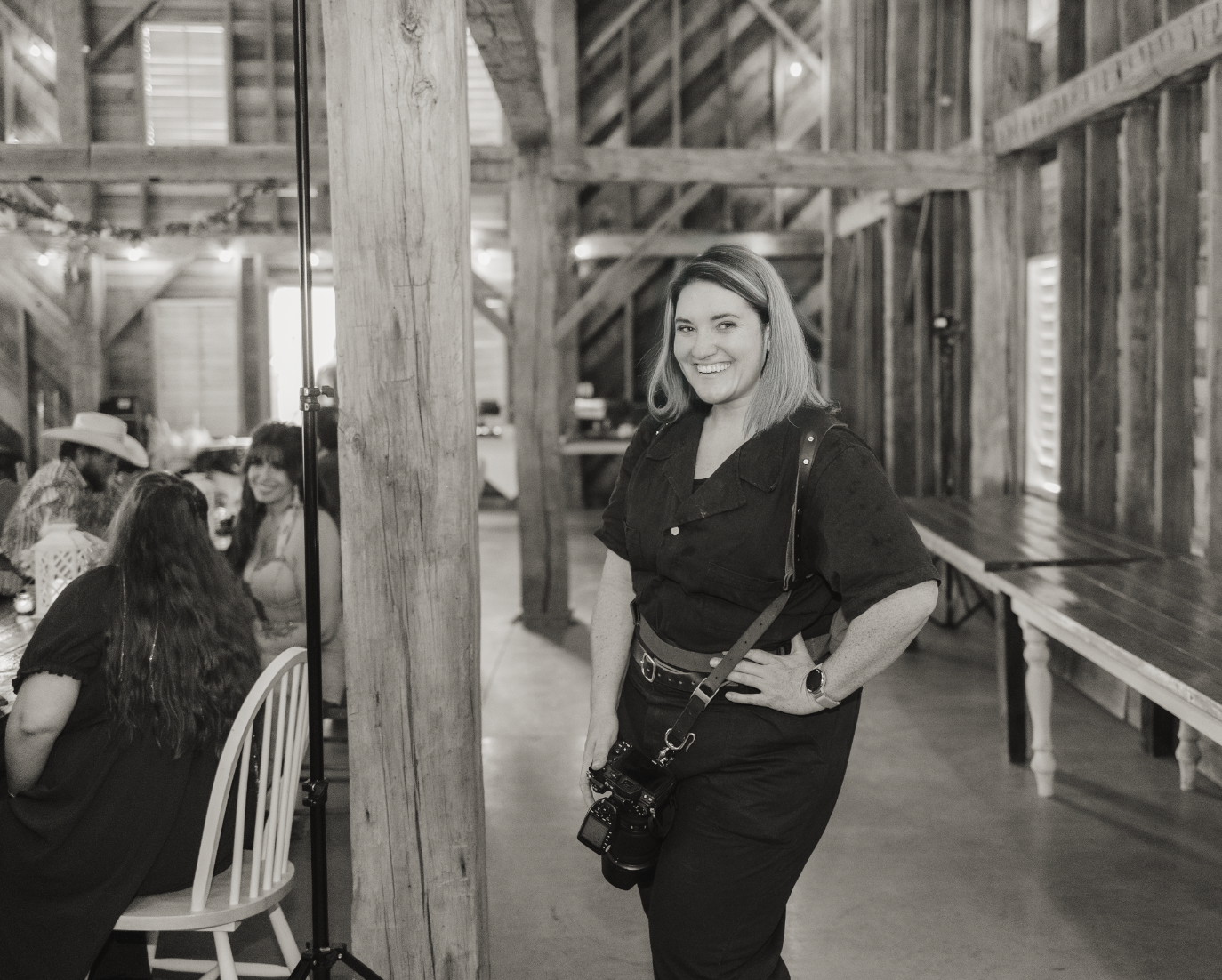 A person in dark clothing poses casually against a wooden post inside a rustic barn-like structure.