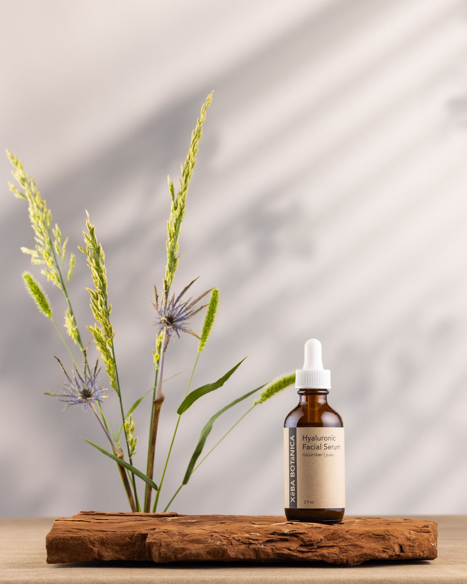 Xeba Botanica Hyaluronic Facial Serum amber dropper bottle beside wildflowers on a wooden surface.