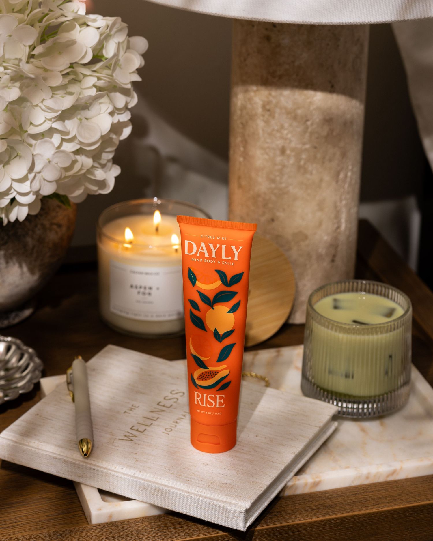 Orange DAYLY RISE skincare tube on marble tray with lit candles, hydrangeas, and a wellness journal.