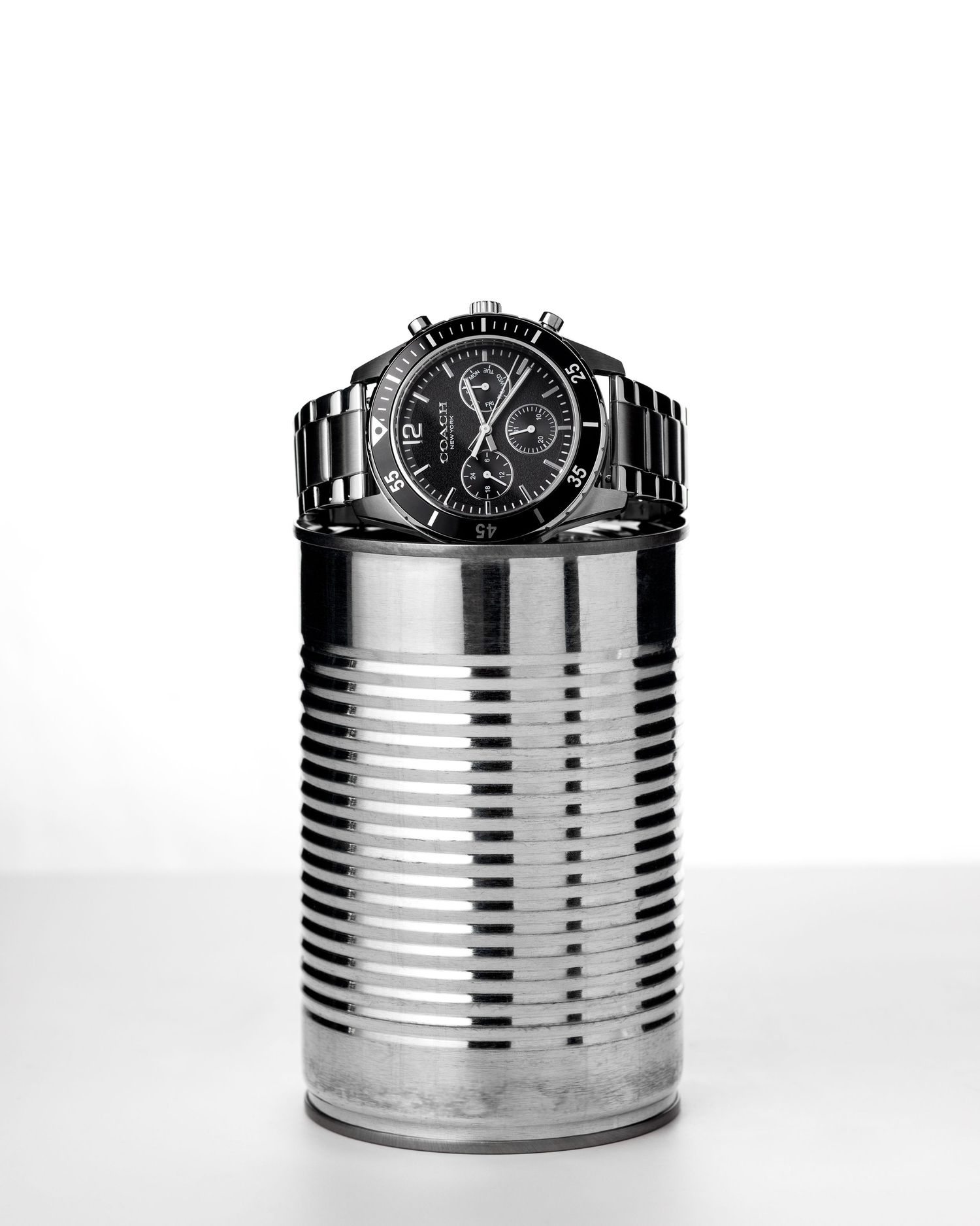 Black chronograph watch balanced on top of a silver tin can against white background.