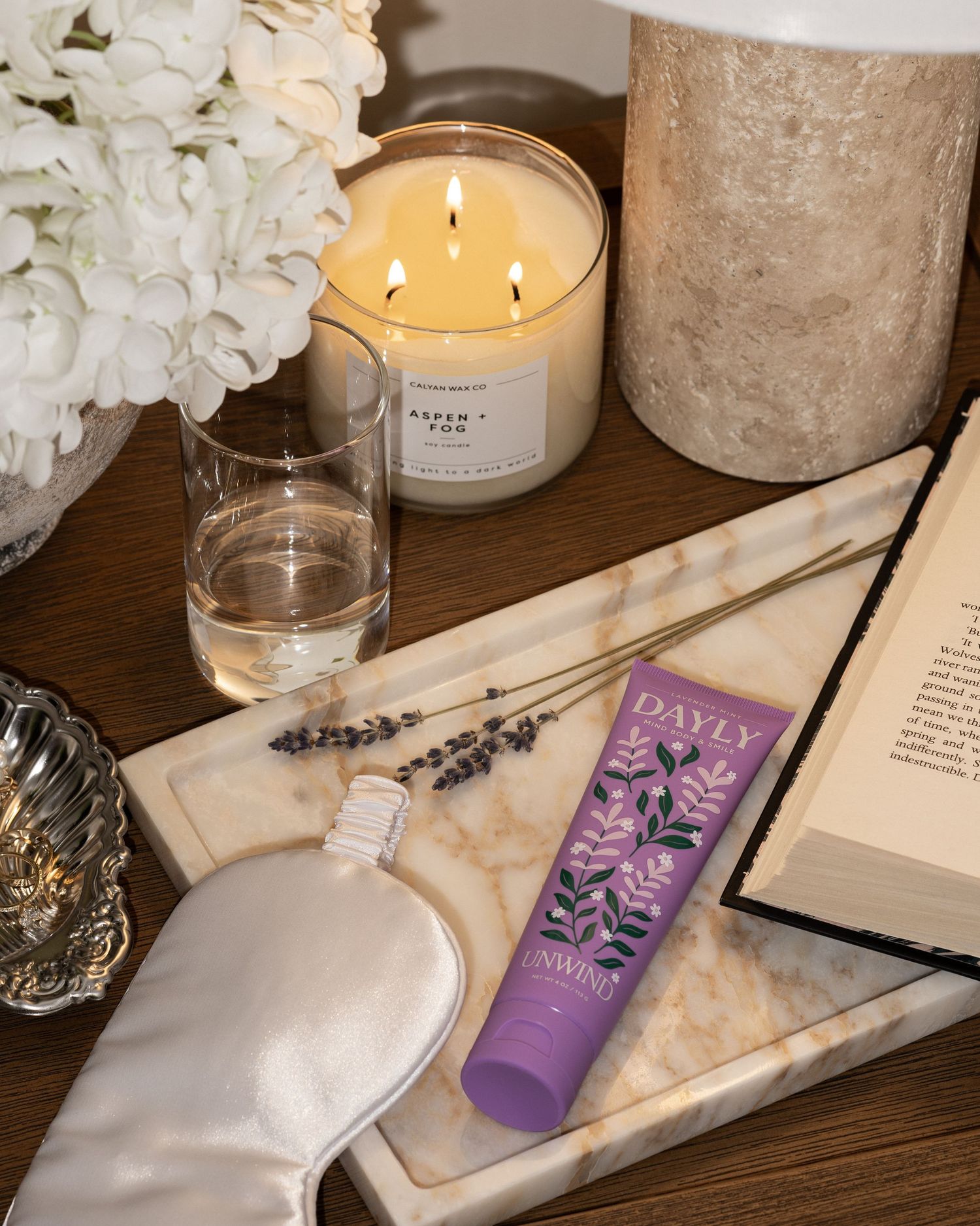 A relaxing bedside tablescape featuring a lit candle, sleep mask, lavender sprigs, and Dayly Unwind hand cream on a marble tray.