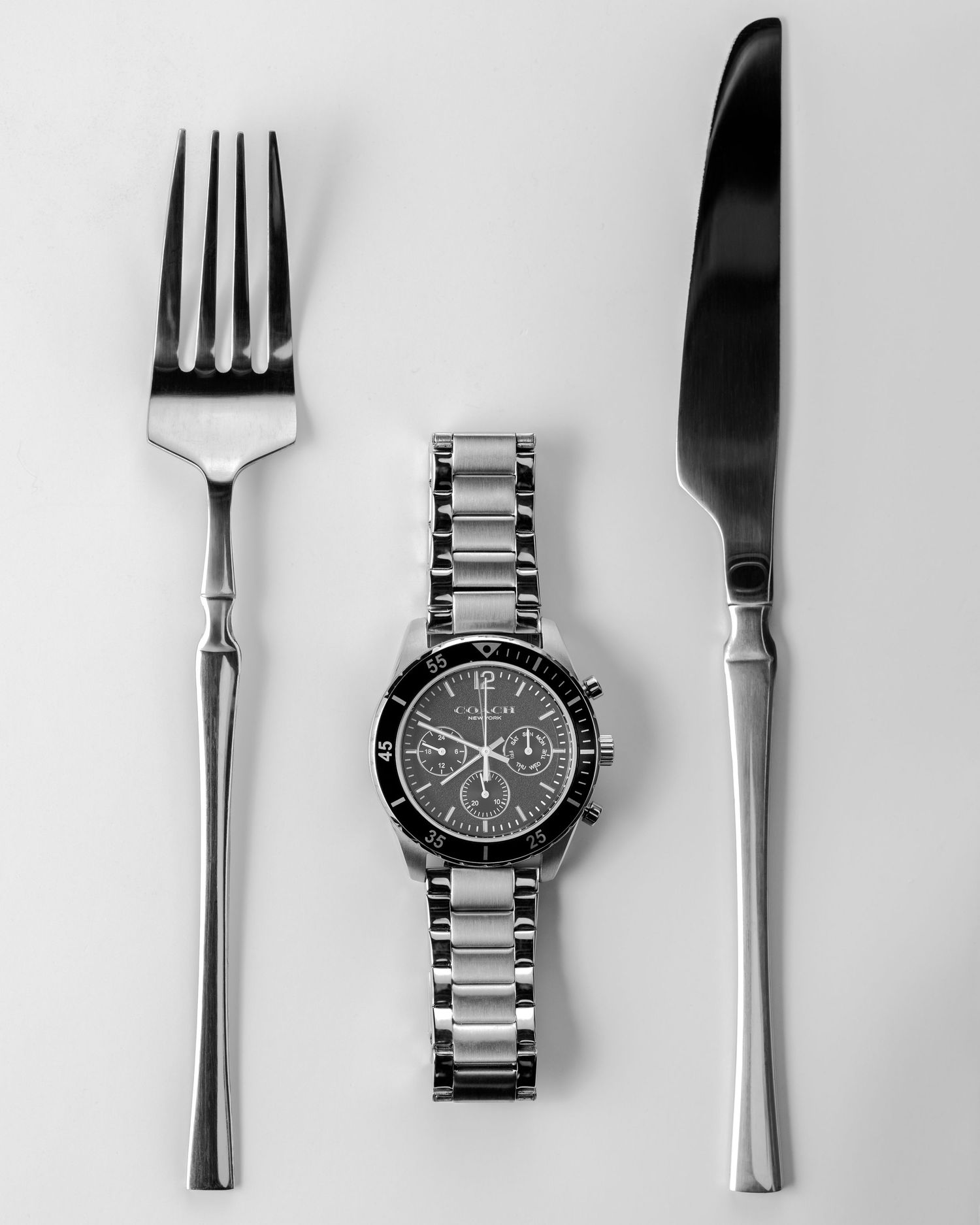 Black and white flatlay of a chronograph watch placed between a fork and knife on white background.