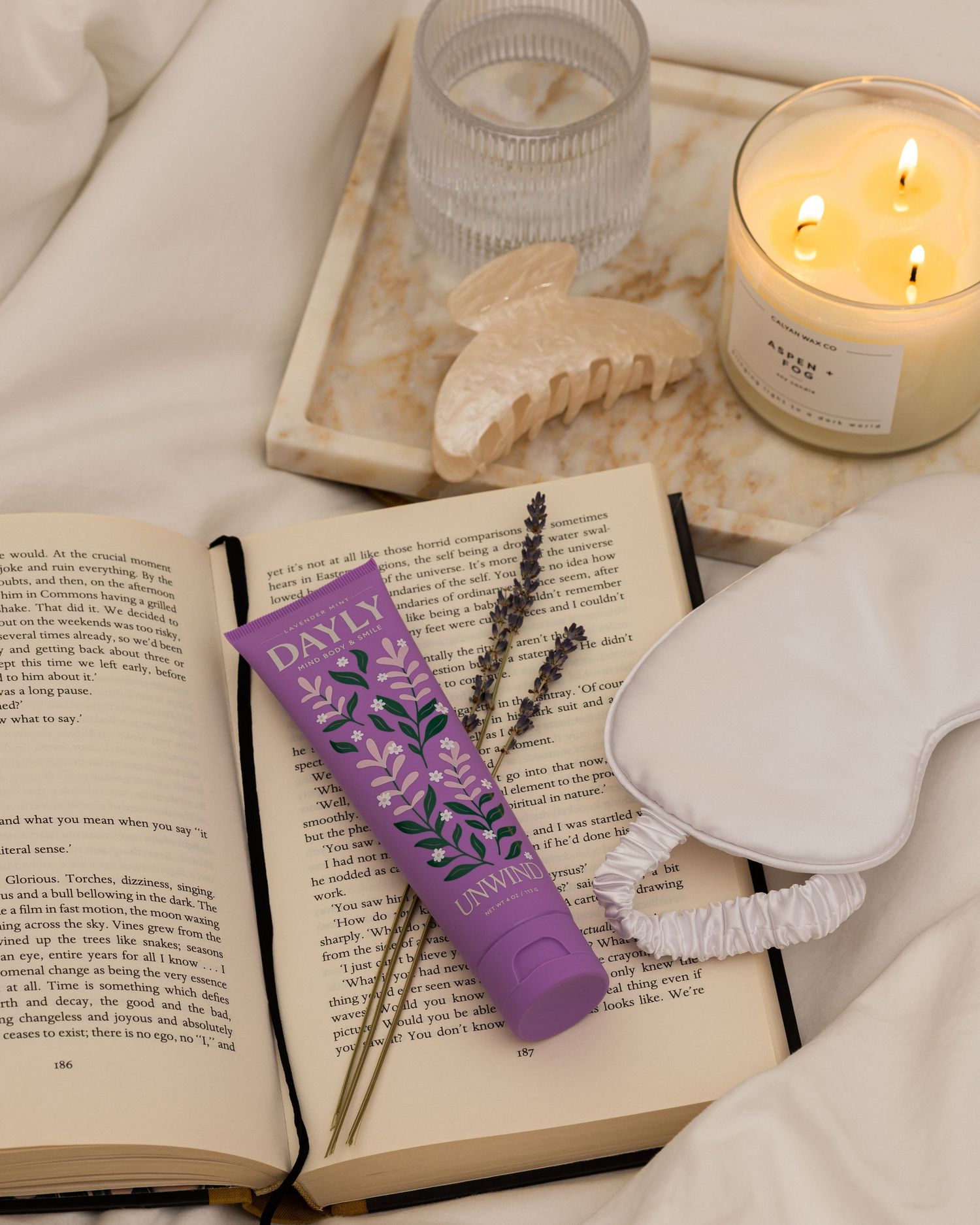 Dayly lavender hand cream on open book with sleep mask, dried lavender, candle, and hair clip on white bedding.