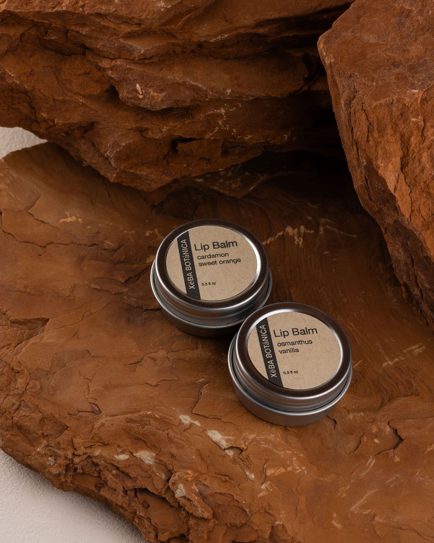 Two XeBA Botanica Lip Balm tins in different scents resting on red sandstone rock surface.