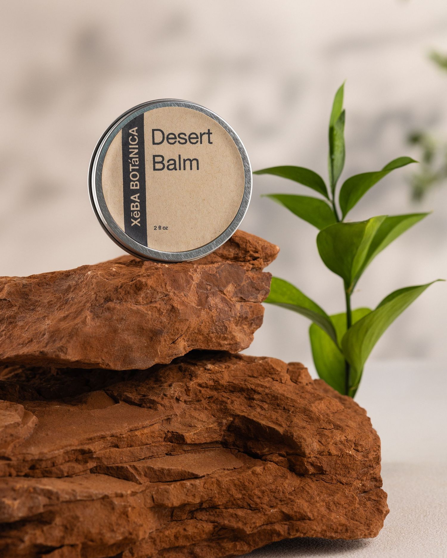 XeBA Botanica Desert Balm tin displayed on red sandstone rocks with green plant leaves in background.