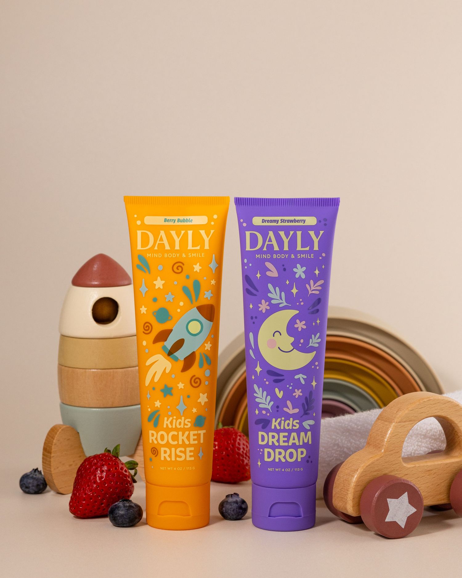 Two DAYLY kids toothpaste tubes, Rocket Rise and Dream Drop, displayed with wooden toys and fresh berries on a beige background.