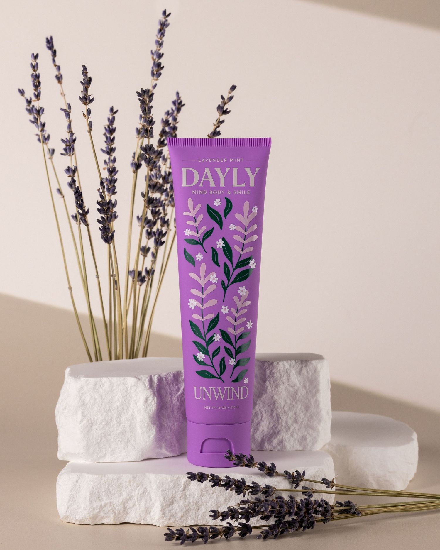 Dayly Unwind lavender body lotion tube in purple packaging displayed on white stones with dried lavender sprigs.