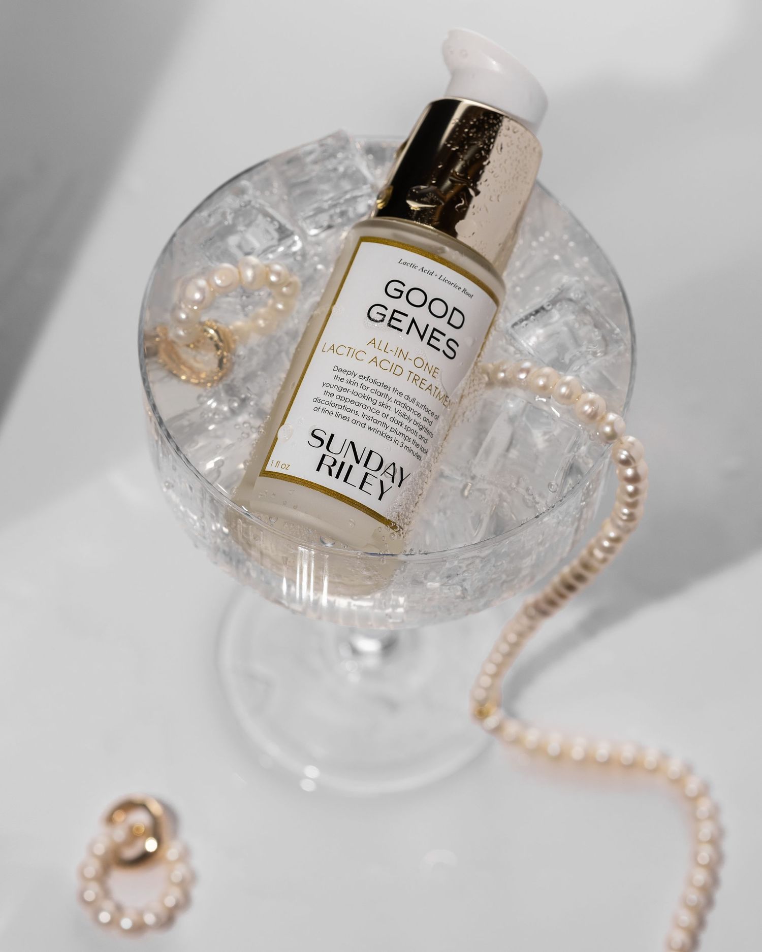 Sunday Riley Good Genes lactic acid treatment bottle resting in a glass of ice with pearl jewelry.