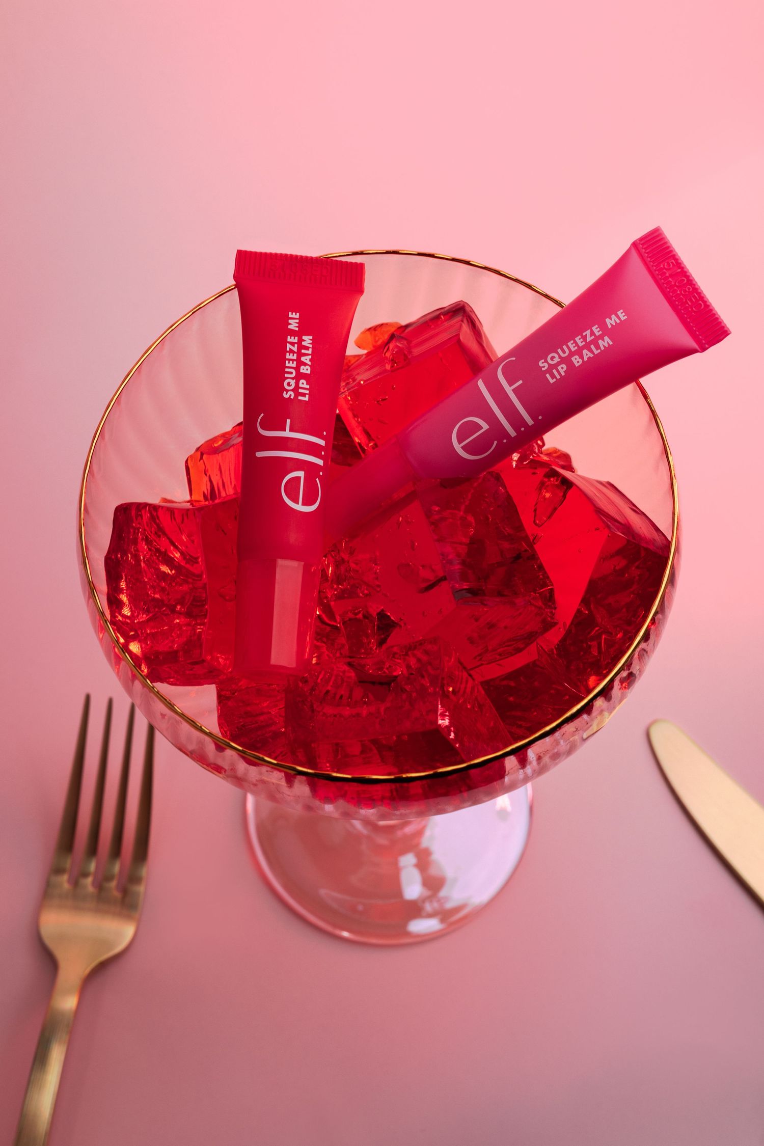 Two e.l.f. Squeeze Me Lip Balm tubes in red and pink resting in a glass bowl of red jello cubes.