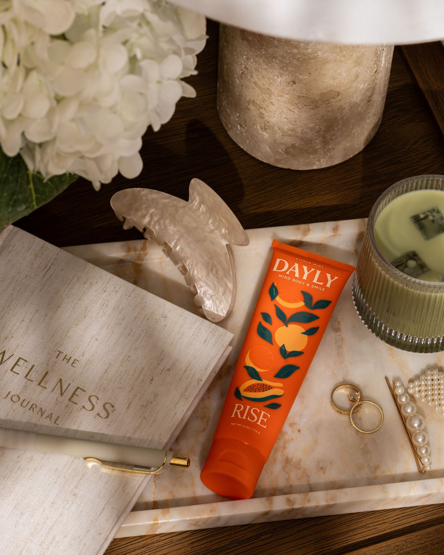 DAYLY Rise orange-scented body lotion tube displayed on marble tray with wellness journal, candle, and white hydrangeas.