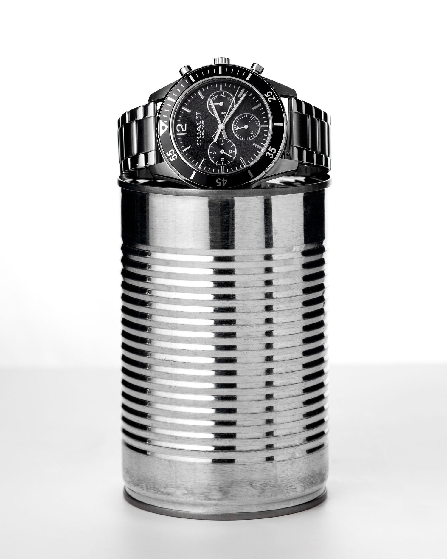 Black chronograph watch displayed on top of a silver ribbed tin can on white background.