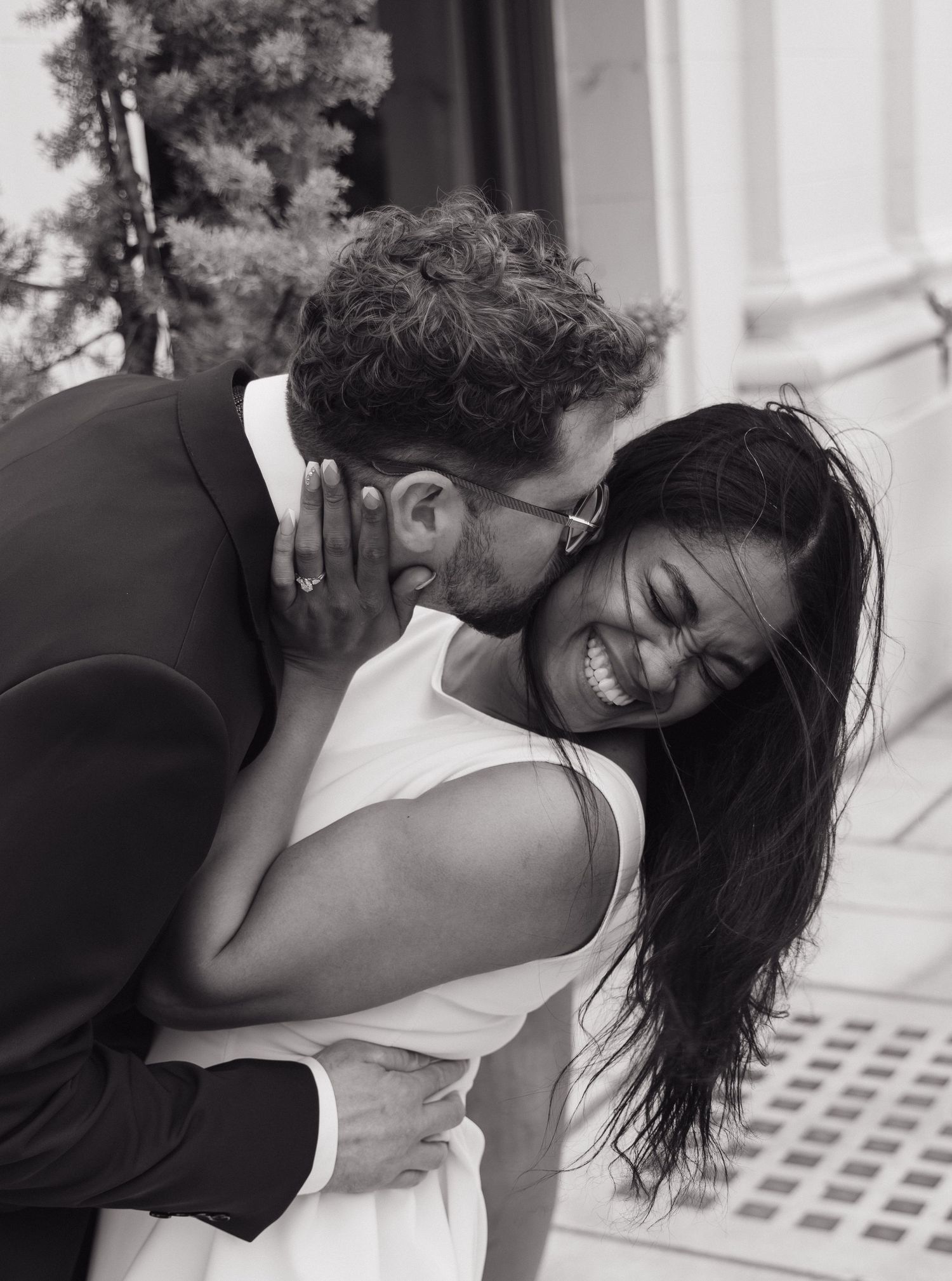 Groom kisses laughing bride on cheek in joyful black and white portrait.