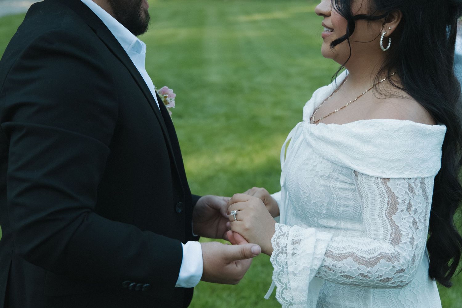 Bride in white lace off-shoulder gown exchanges rings with groom in black suit outdoors on green lawn.