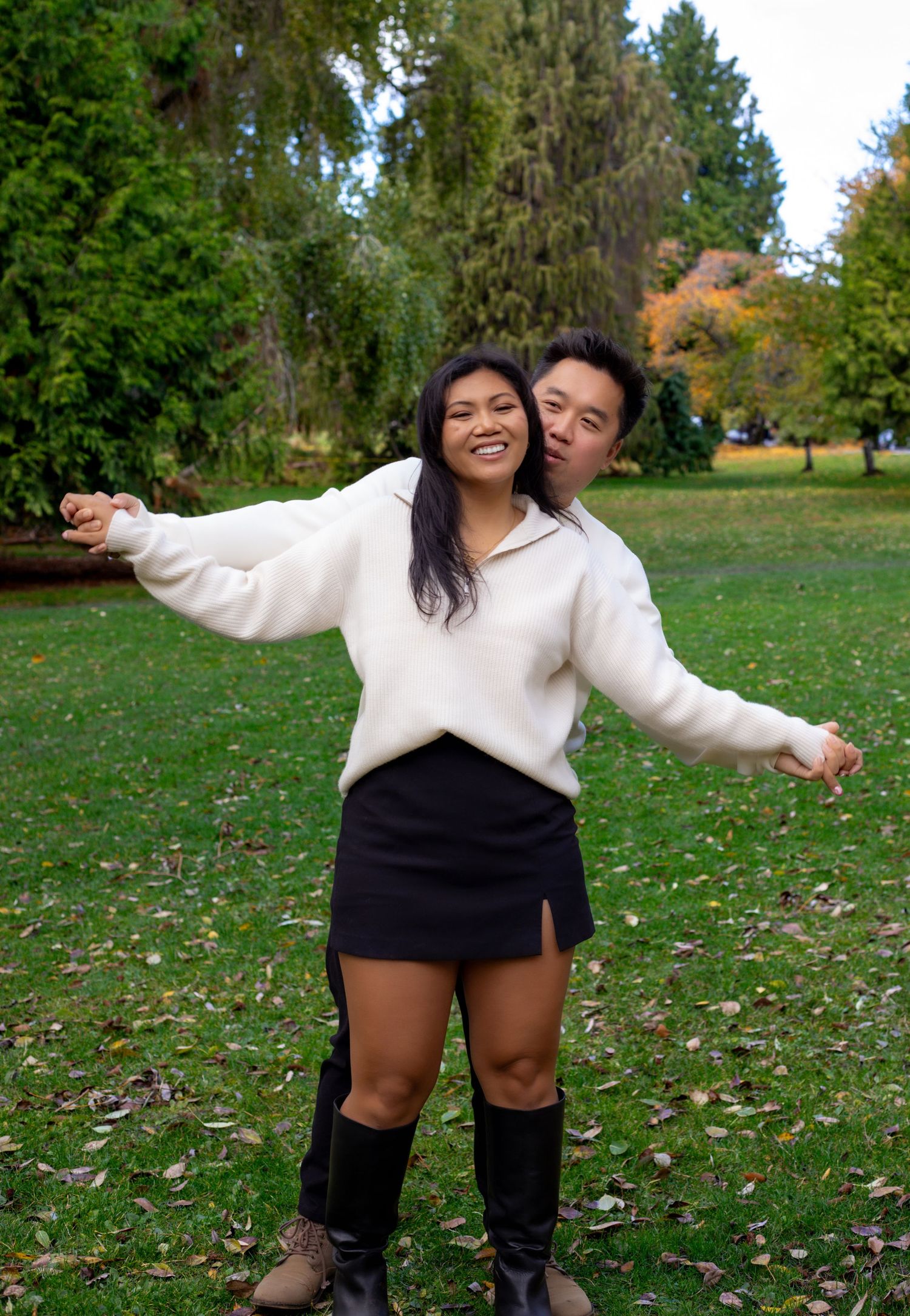 A smiling couple poses playfully in a park, woman in white sweater and black mini skirt, man hugging her from behind.
