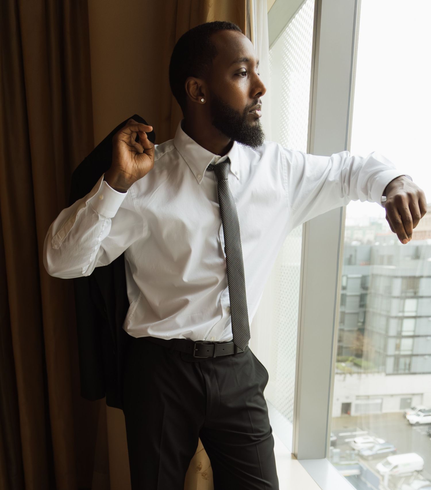 Stylish groom in white shirt and tie holding jacket while gazing out hotel window.