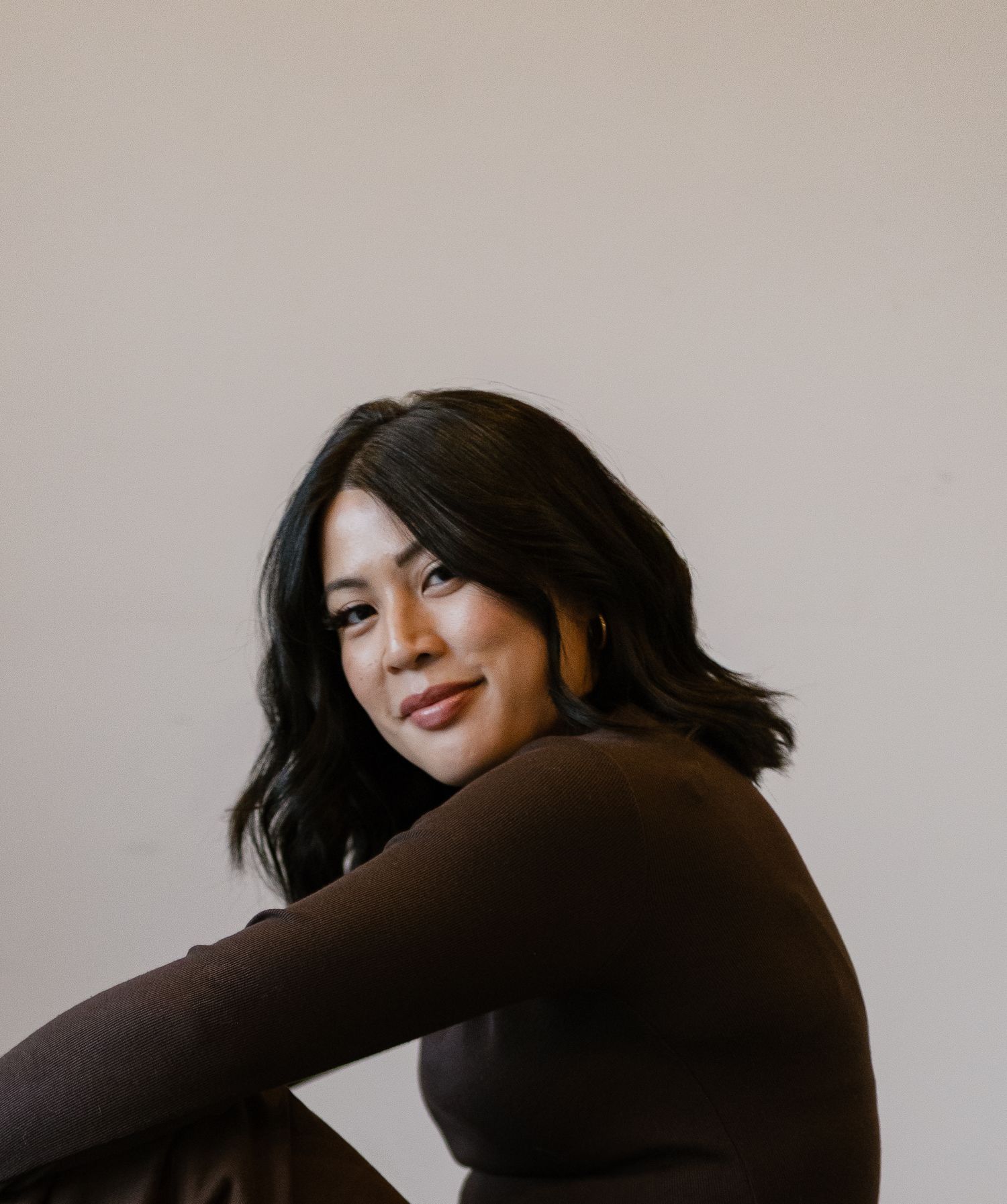 Smiling woman in brown turtleneck with dark wavy hair poses casually against light background.