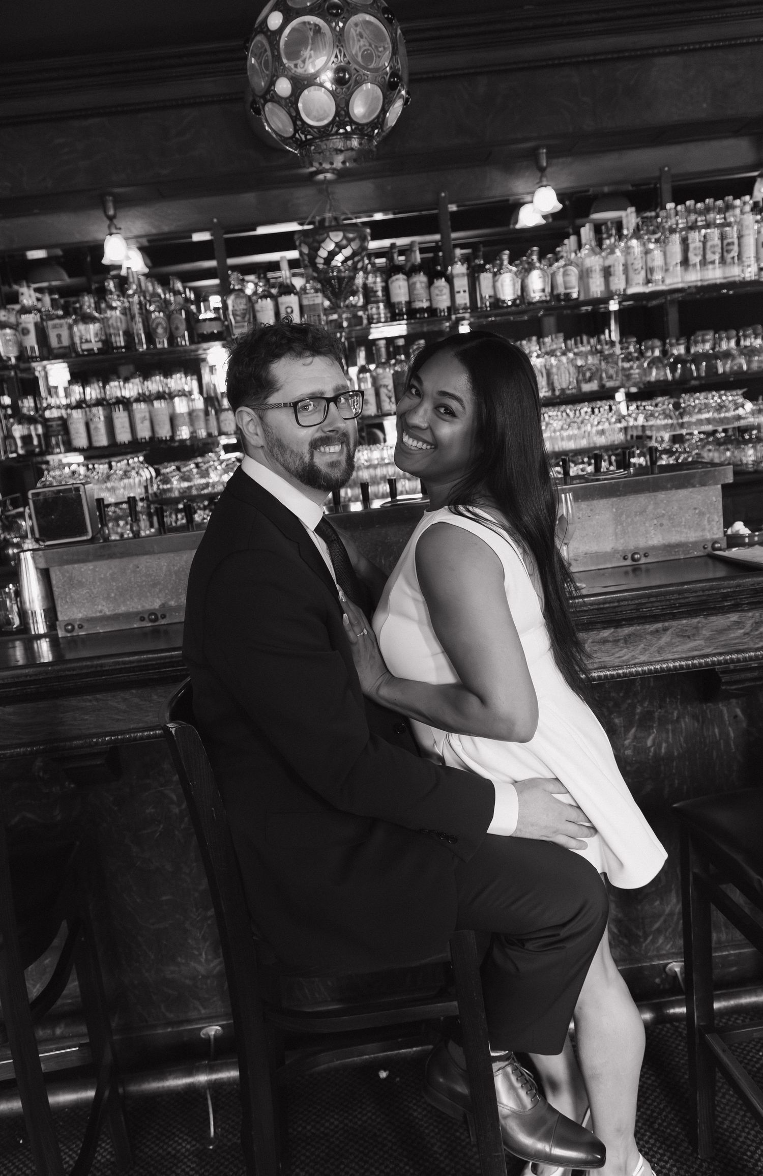 Black and white photo of smiling interracial couple posing together on bar stools at a bar.