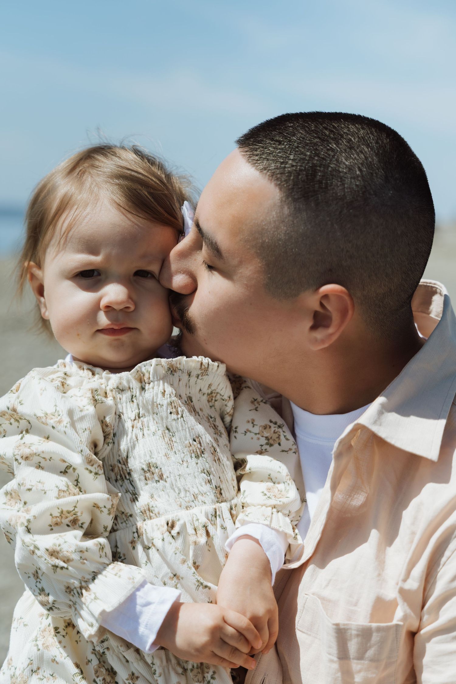 A father tenderly kisses his baby daughter on the cheek outdoors near the beach on a sunny day.