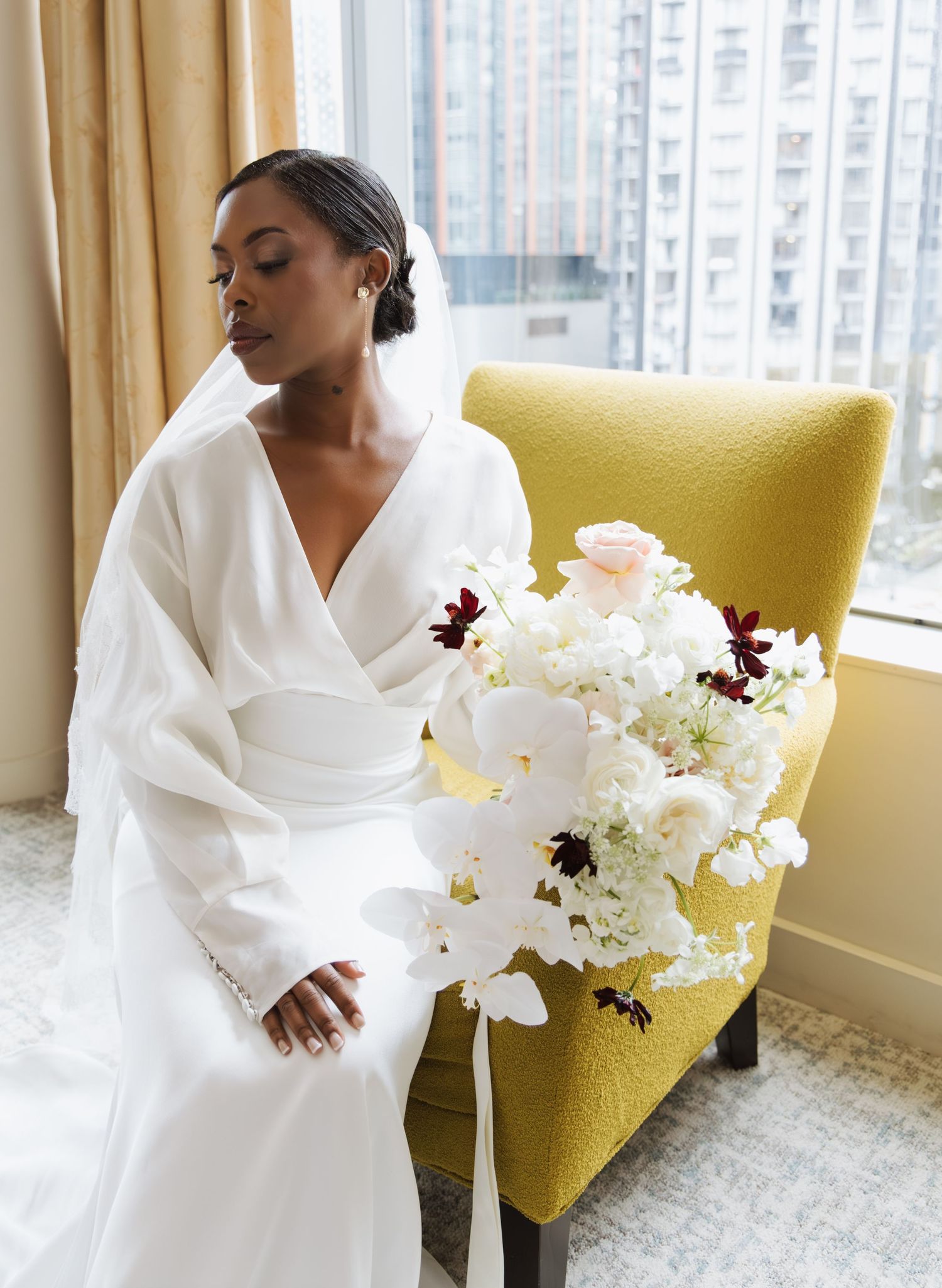 Elegant bride in white wrap wedding dress holds lush white and burgundy bouquet seated in gold velvet chair.