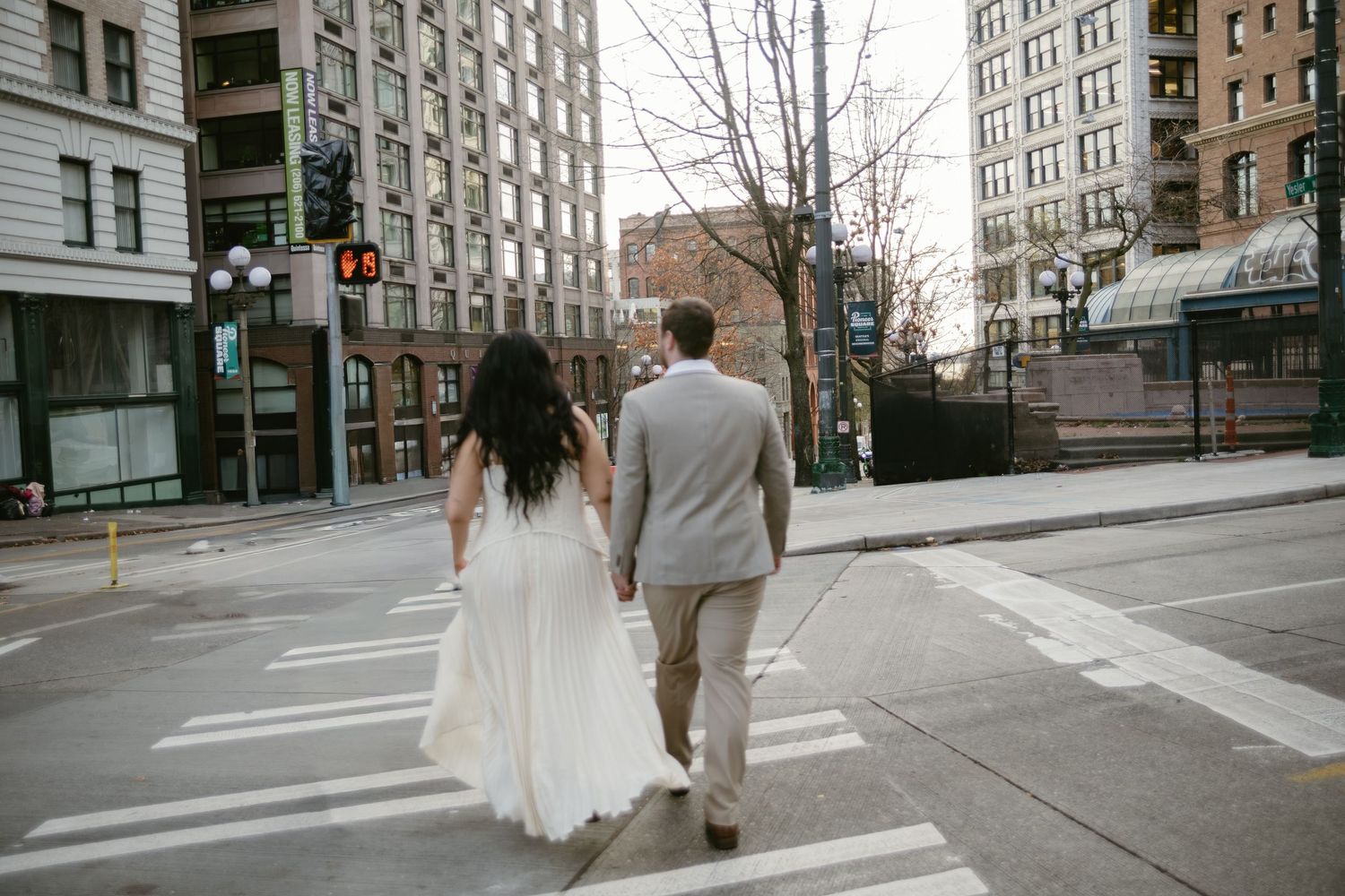 Bride and groom walk hand-in-hand across urban crosswalk in city wedding photo.