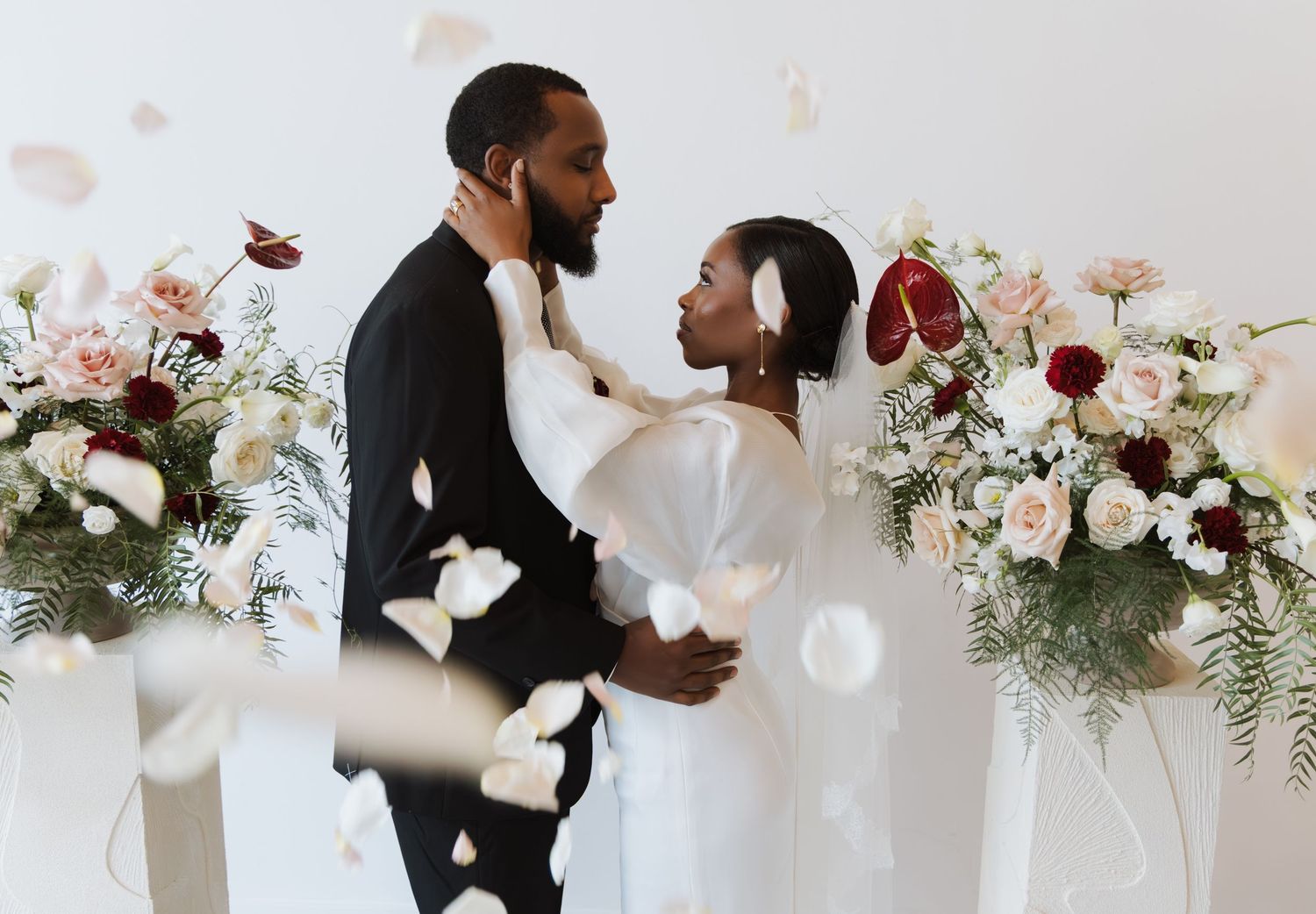 Black bride and groom embracing at altar with pink and burgundy floral arrangements, petals falling around them.