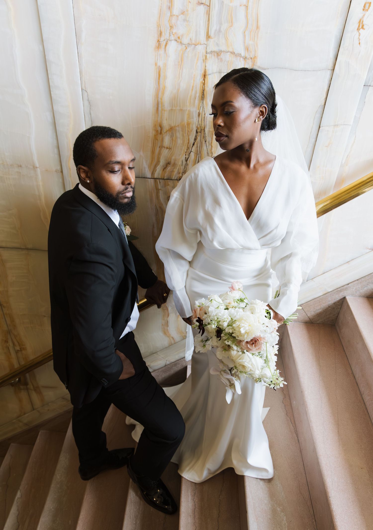 Bride and groom in formal wedding attire pose on marble staircase, bride holding white floral bouquet.